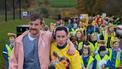 Two men walking in front of a large crowd of primary school children.