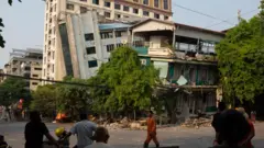 Man in orange outfit walks in front of a damaged building, tipped on its side with a damaged tree and telephone wires.