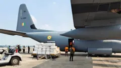 A military plane parked at an airport, with people on hand unloading cargo