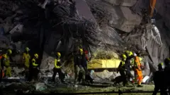 Crew of people in yellow outfits and hard hats walking in front of a building reduced to rubble in Bangkok. It is dark with a spotlight shining on them.