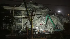 A building destroyed by an earthquake. There is a green digger in front of it along with a group of rescue workers.
