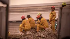 Five rescue workers in yellow safety suits wait on top of a pile of rubble