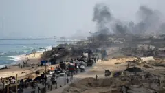 A long line of people and cars carrying belongings along a road out of part of Gaza City, which is smoking and in ruins