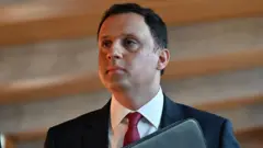 A man in his 40s is wearing a black suit and red tie as he is holding a black case.