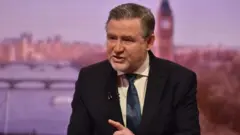 Barry Gardiner wearing a black suit jacket and blue tie and sitting in front of a backdrop of the London skyline