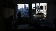 A man stands in a dark room and stares out at a pile of rubble