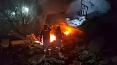 Two men look on as fire burns under a destroyed building surrounded by rubble