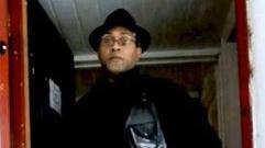 An old scanned image of Louis Campbell. He is wearing a black leather jacket, black polo top and a black hat. he has clear framed glasses and is standing in a wooden doorway.