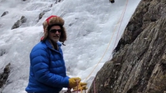 Man wearing a red helmet, blue puffer jacket and yellow gloves climbing an icy rock face, secured by ropes.