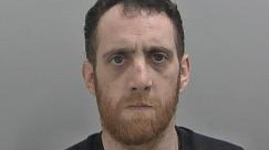 Mugshot of a man in a black top, stood in front of a grey background