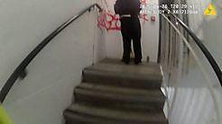 A still image from a police body-worn camera, showing a flight of stairs, and at the top of the stairs, a person wearing black with their back to the camera, and some red graffiti on the white wall.
