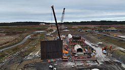 The image is taken from the air by a drone.  It shows a large construction site, where a new reservoir is being built.  There is a crane in the background, and another crane in the foreground, which is in the process of lifting a large brown steel panel, which is a new wall at the reservoir.  To the right there is a concrete tunnel and a trench where the wall will be lowered into place.