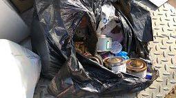 An open black bin liner with rubbish spilling out into the street including cans