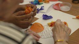 Close-up of two pairs of hands working on a collage - one pair is that of an old person, the other of a younger care worker. This shot was taken in June 2025.