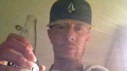 Christopher Oates, wearing a baseball cap, holds a beer bottle up to the camera.