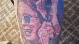 Tattoo of Ozzy Osbourne which is him smiling at a microphone stand with round sunglasses on. 