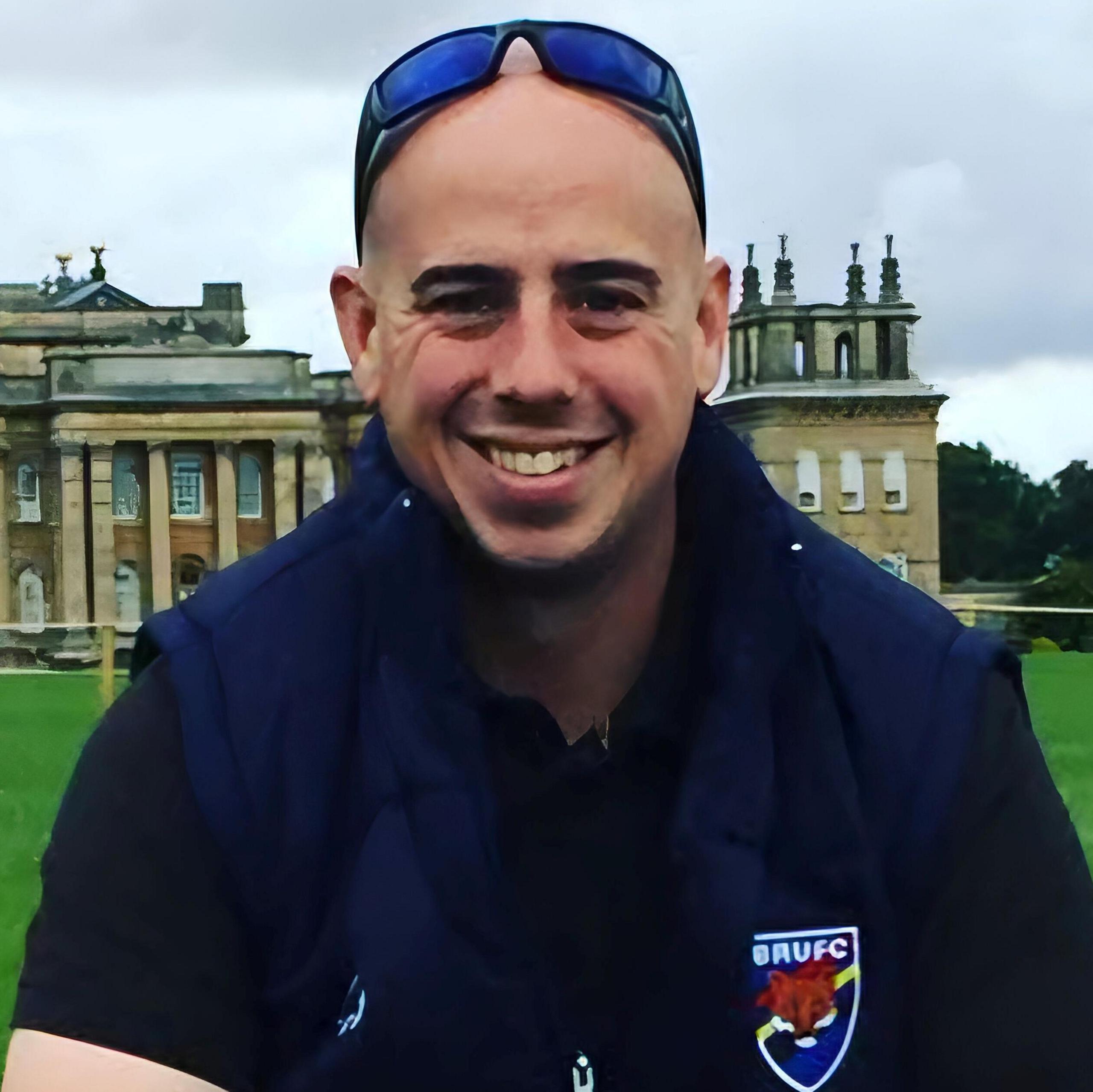 Martyn Sadler with stately home behind him - he is wearing a dark blue rugby top and has sunglasses on his head. He is looking directly at the camera and smiling.