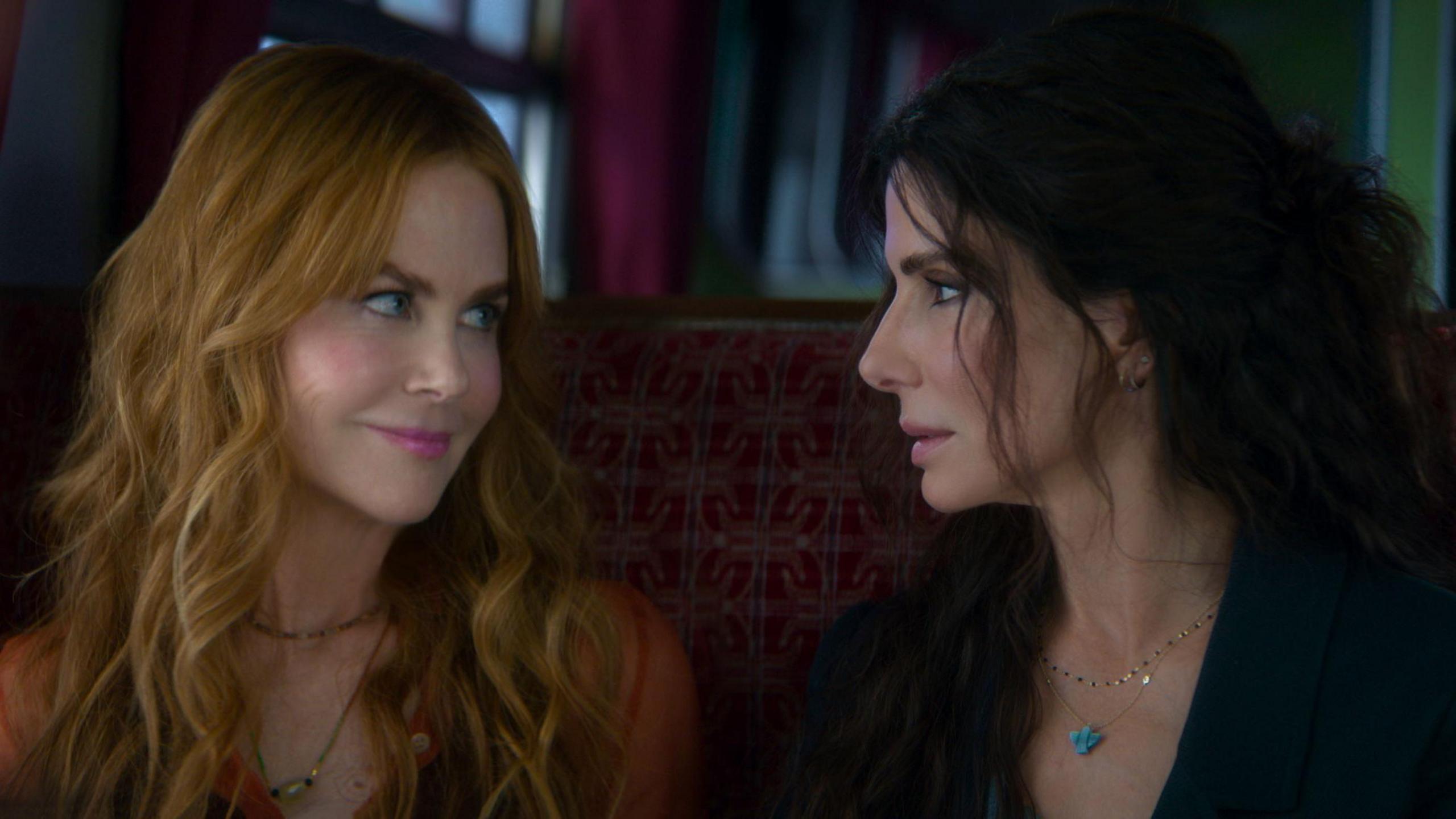 Nicole Kidman, with red long wavy hair, smiles at Sandra Bullock, who has dark long hair pulled back off her face, and are sitting in a train carriage.