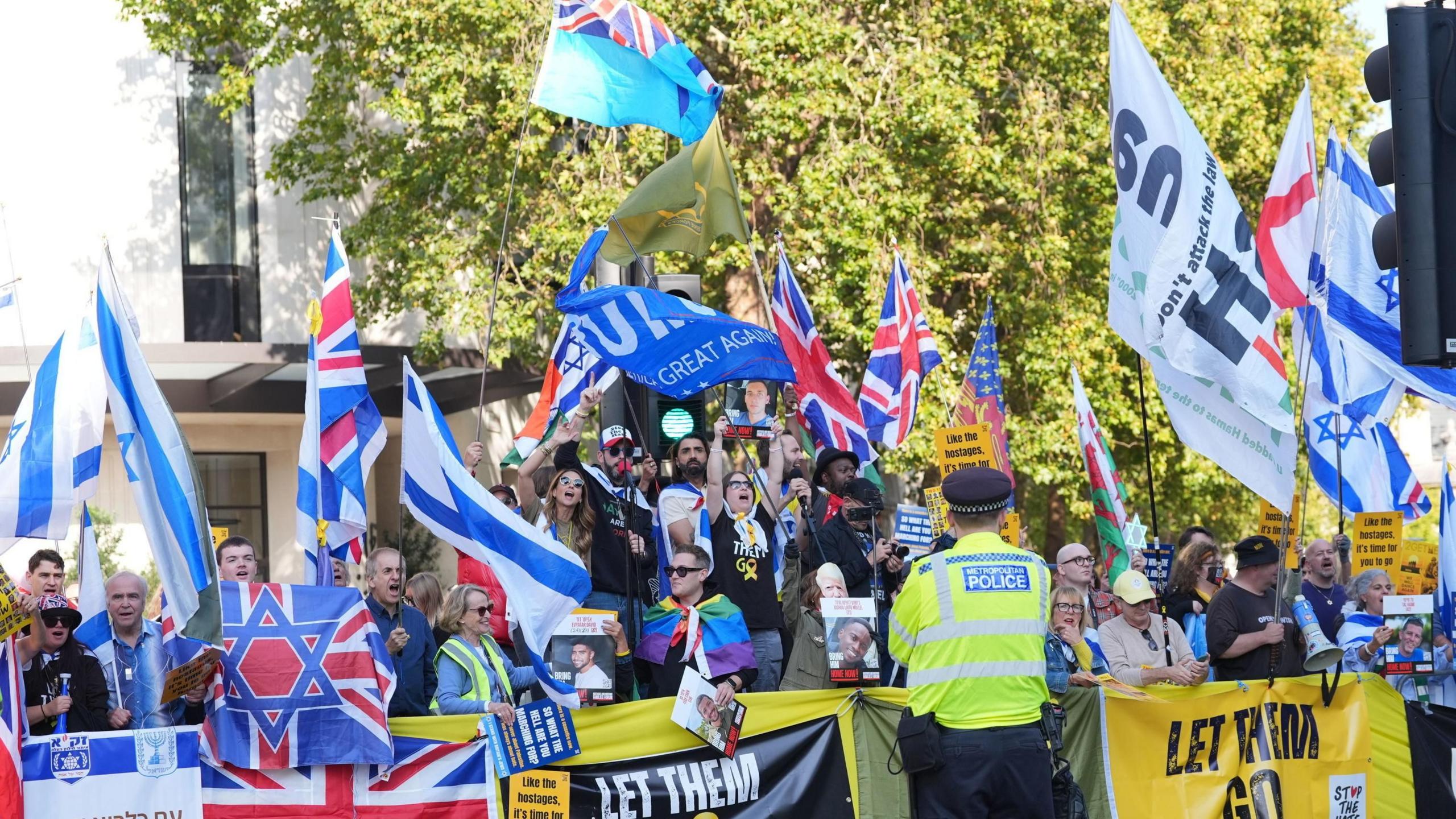 A crowd waving various flags stands behind a low fence row of banners, as a police officer with his back to the camera watches on. Many hold Israeli flags aloft, while some union jacks and St George's flags can also be seen. The crowd consists of men and women, and some are shouting.