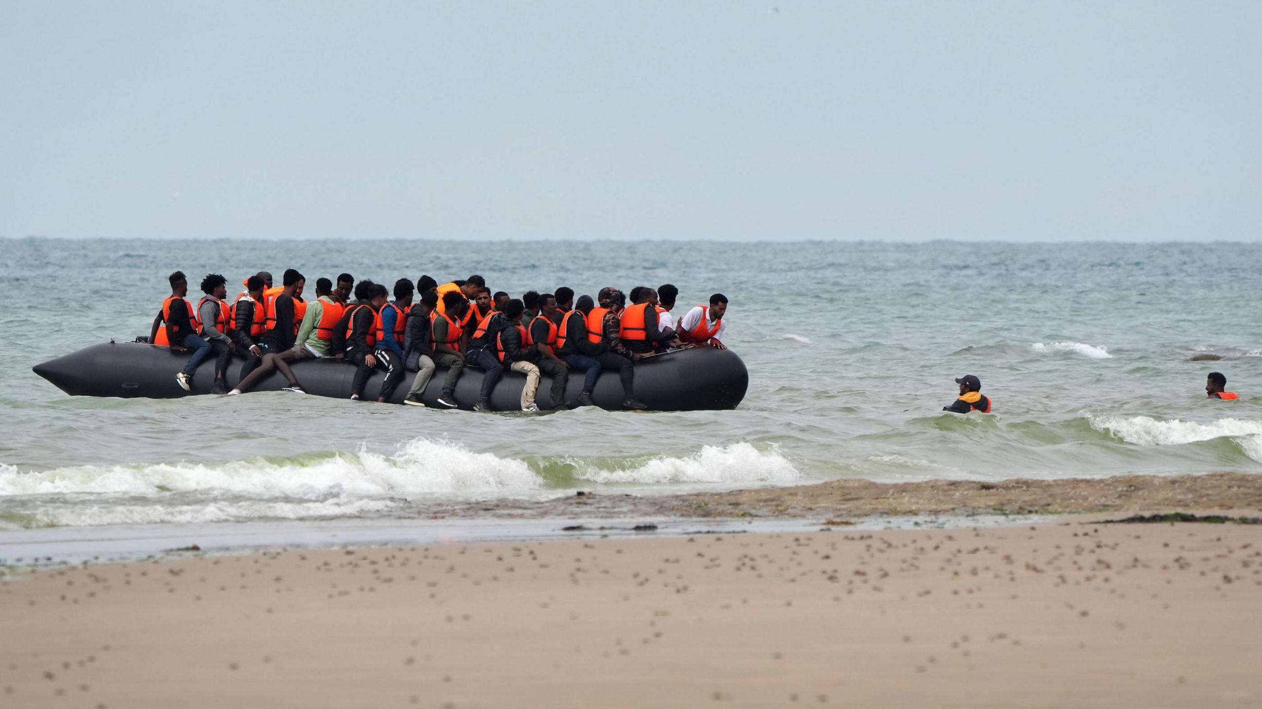 Near the shore at Wimereux, France, a group of people believed to be migrants, wearing orange life jackets, board an overcrowded inflatable boat. Some are already seated while others are still in the water trying to climb aboard. Waves crash onto the beach in the background.
