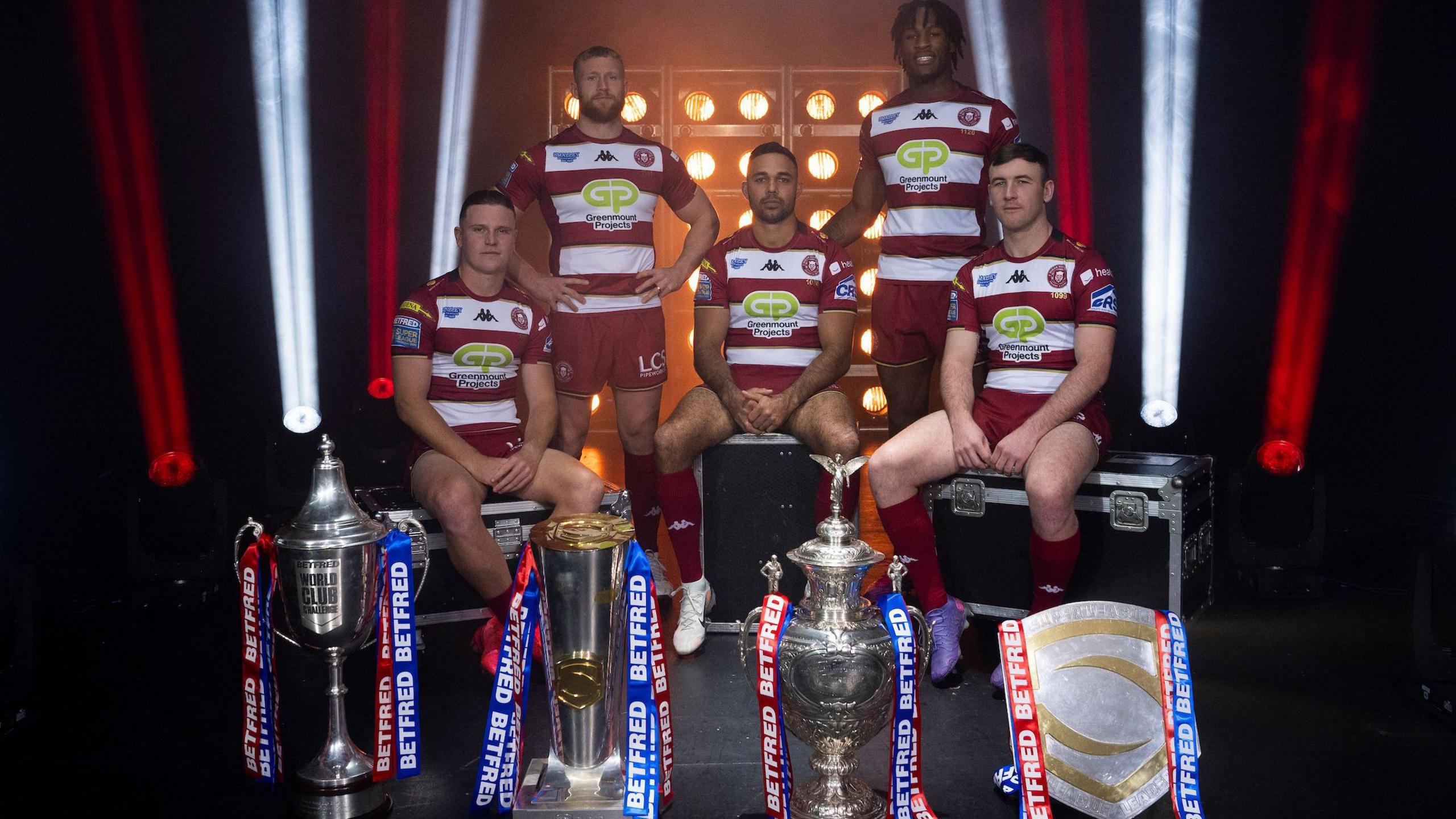 Wigan players Jai Field, Luke Thompson, Bevan French, Junior Nsemba and Harry Smith sit behind their four 2024-won trophies in the World Club Challenge, Super League, Challenge Cup and league leaders shield for a promotional photograph in a red and white lit studio