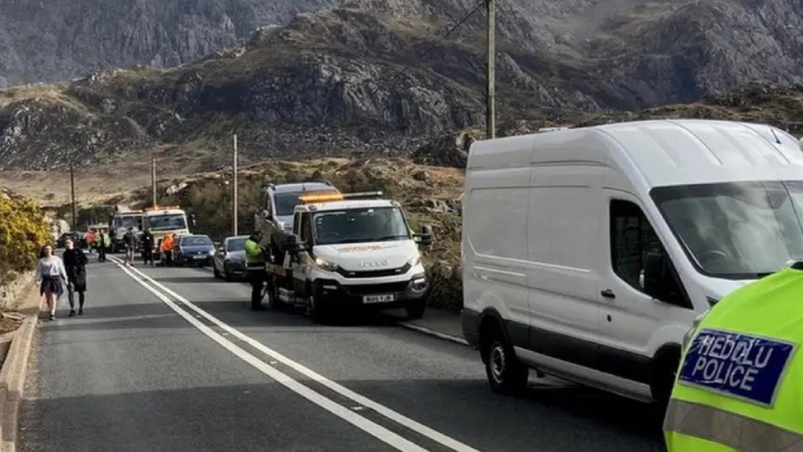 The back of a police officer (right) in high vis uniform and hat standing on the A5 in the area of Llyn Ogwen, several vans line the road, including tow trucks taking cars away. The mountains of Eryri cut through the sky in the background. 