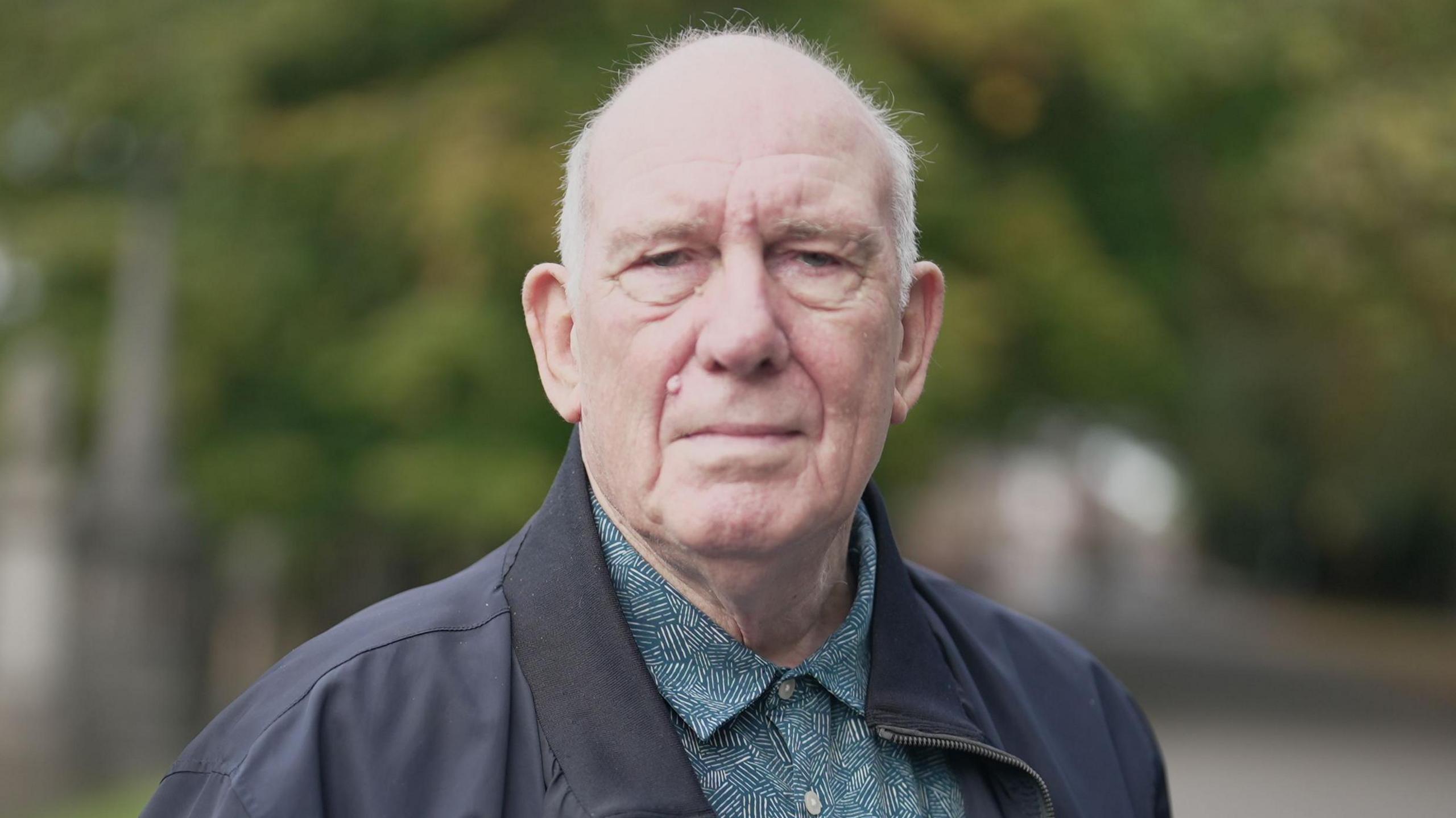 Roy Pollitt, wearing a green and white patterned shirt and dark jacket, is photographed in a park.