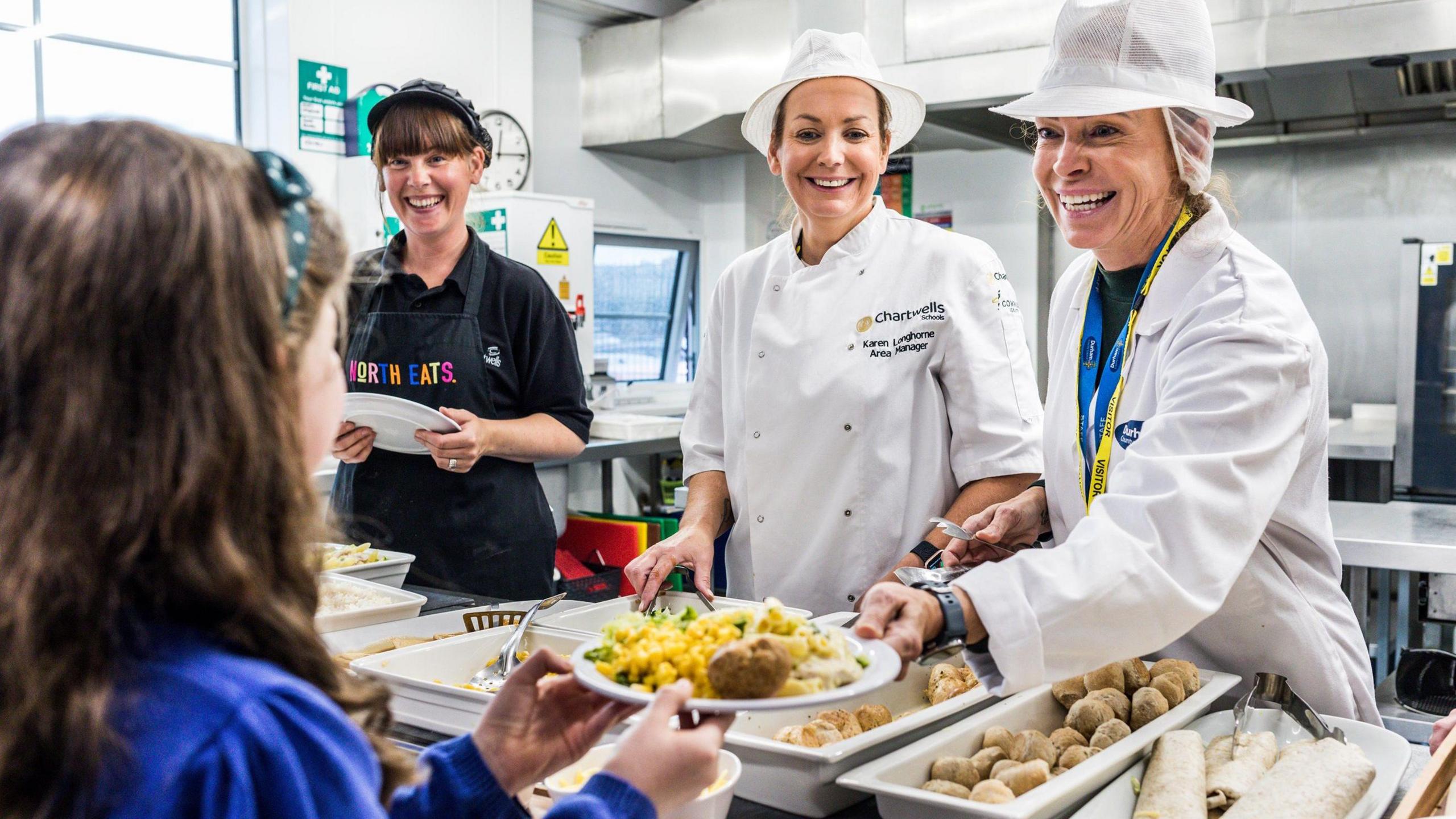 A young school girl is being handed a plate of food from a serving counter by three smiling dinner ladies. She is turned away from the camera but has long brown hair and a blue school jumper. Two of the three dinner ladies wear white overalls and hair nets, while the third has a black apron on.