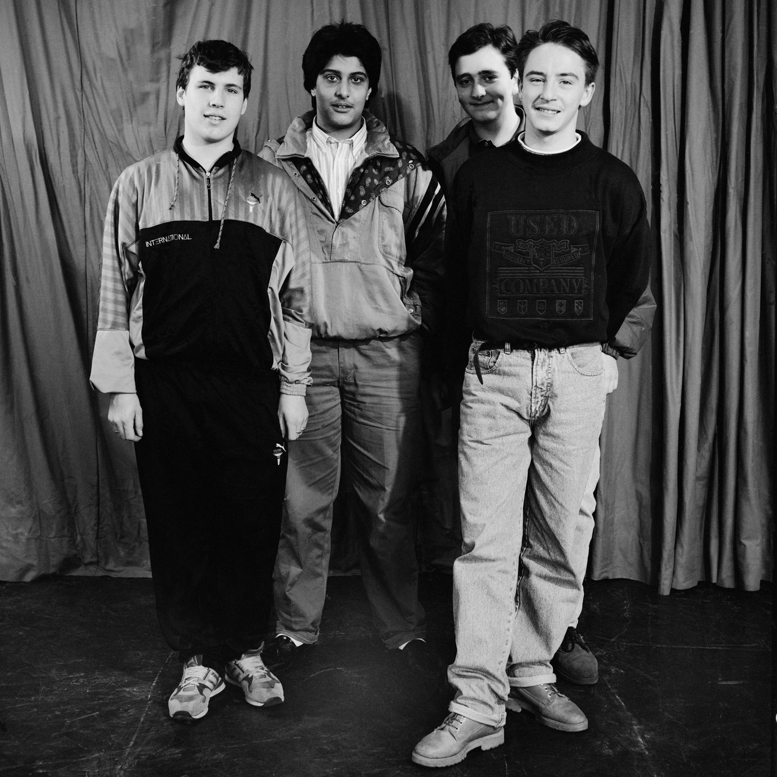 Boys from Tulse Hill School, 1990. Four young men stand in front of a curtain. They wear jeans or tracksuit bottoms, and casual tops. Black and white photo.