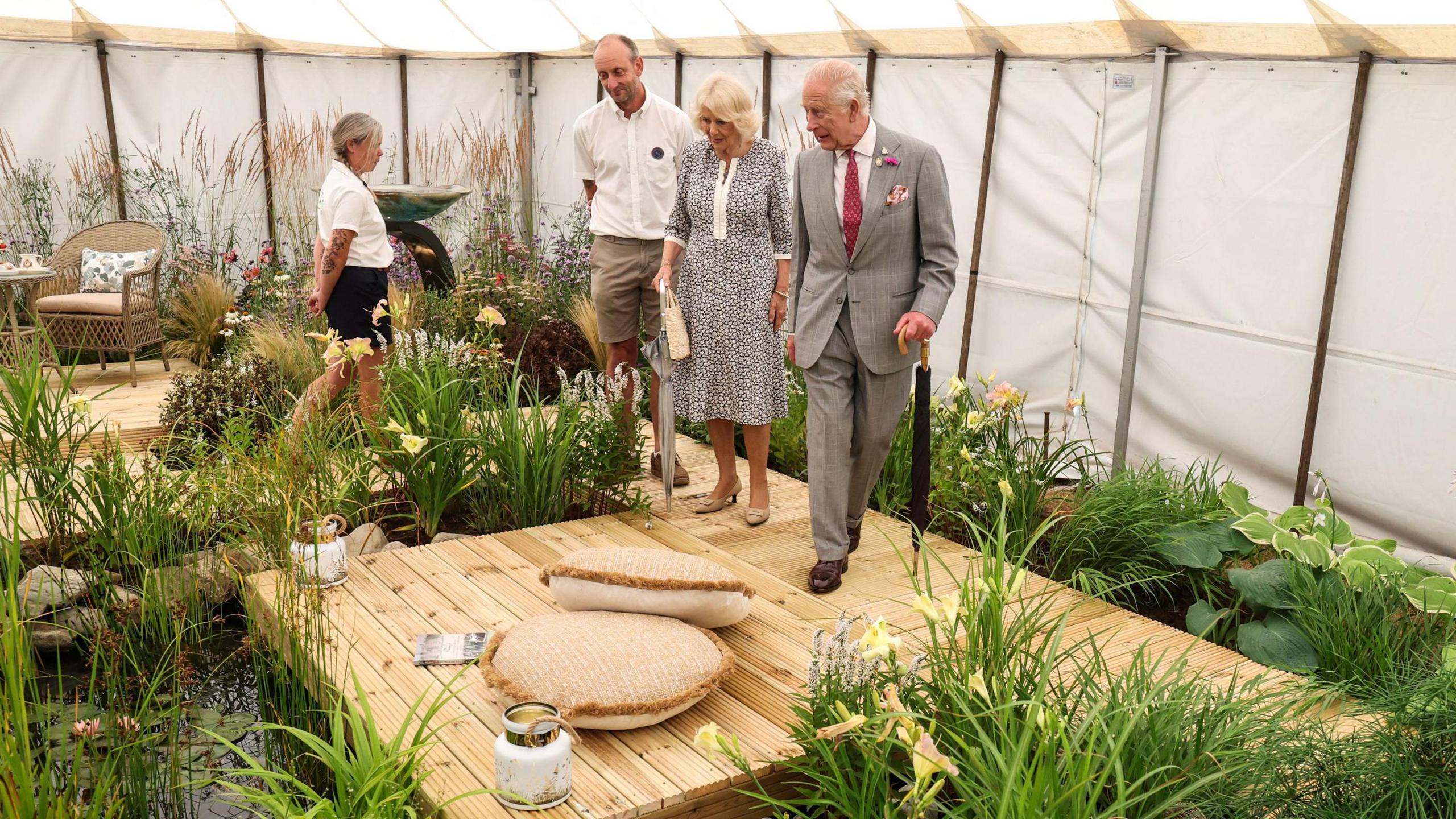 King Charles III walking through an indoor garden display at the Sandringham Flower Show with Queen Camilla walking behind him. They are walking across a wooden plinth. King Charles III is dressed a pale grey suit with a burgundy coloured tie. Queen Camilla is wearing a black and white floral dress. A man and woman are following behind them. 
