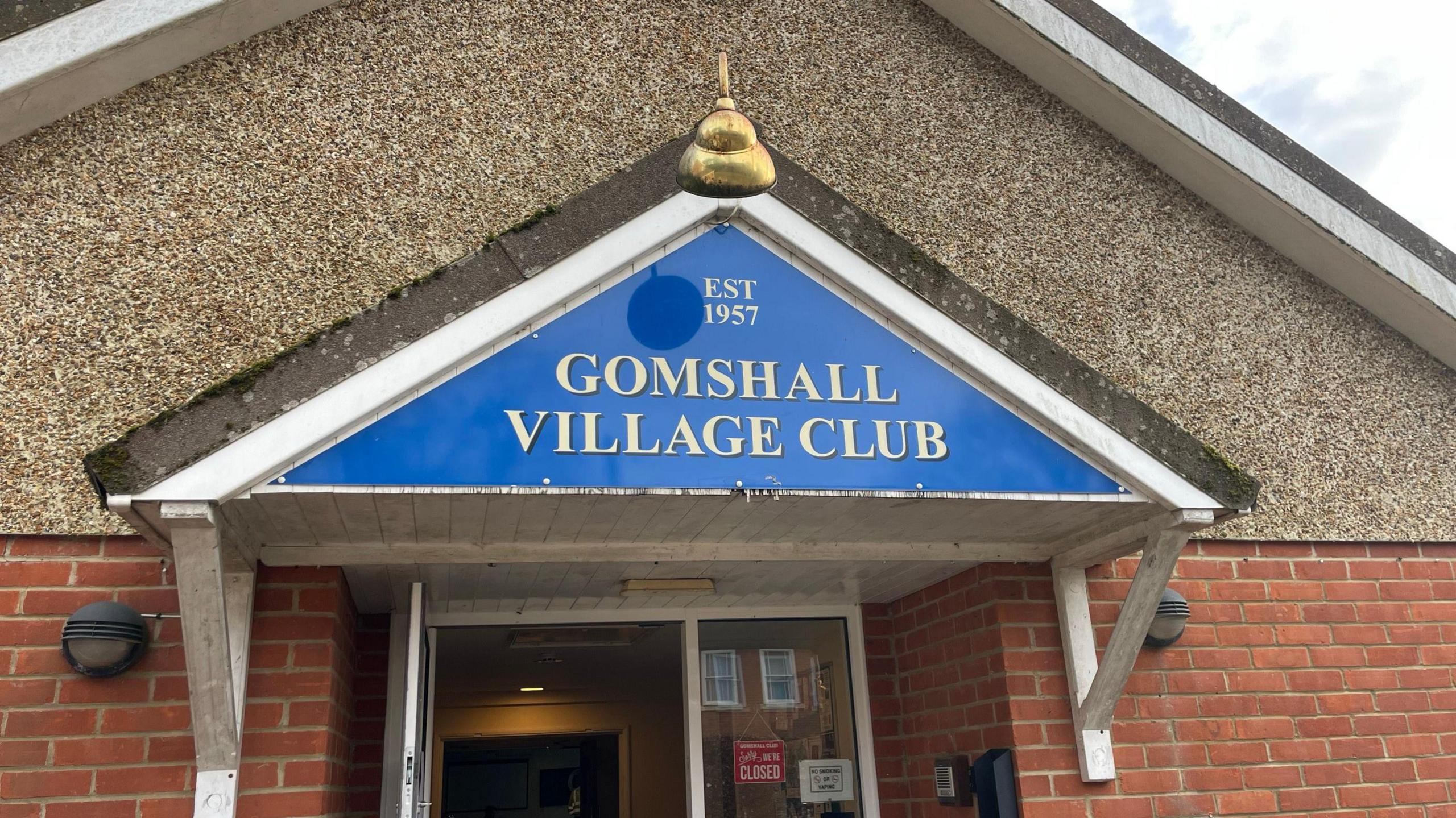 The entrance to Gomshall Village Sports & Social Club. Above the door is a sig with a blue background and gold writing saying "Gomshall Village Club"