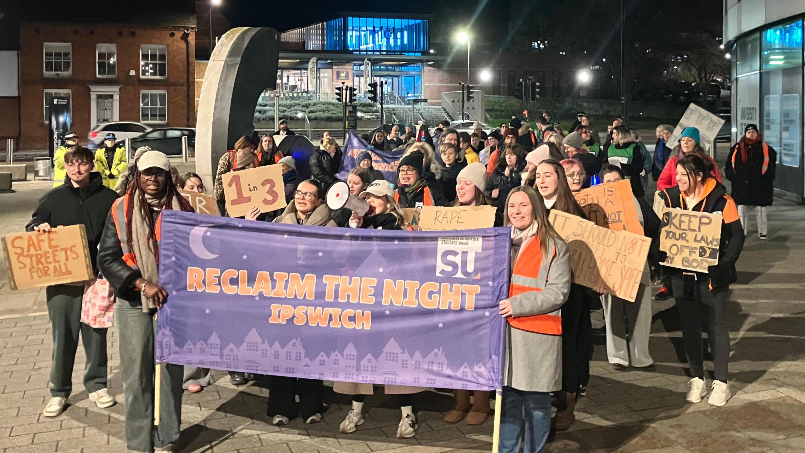 A large crowd of people are stood outside. It is dark. There is a big purple banner at the front which reads: Reclaim the Night Ipswich.