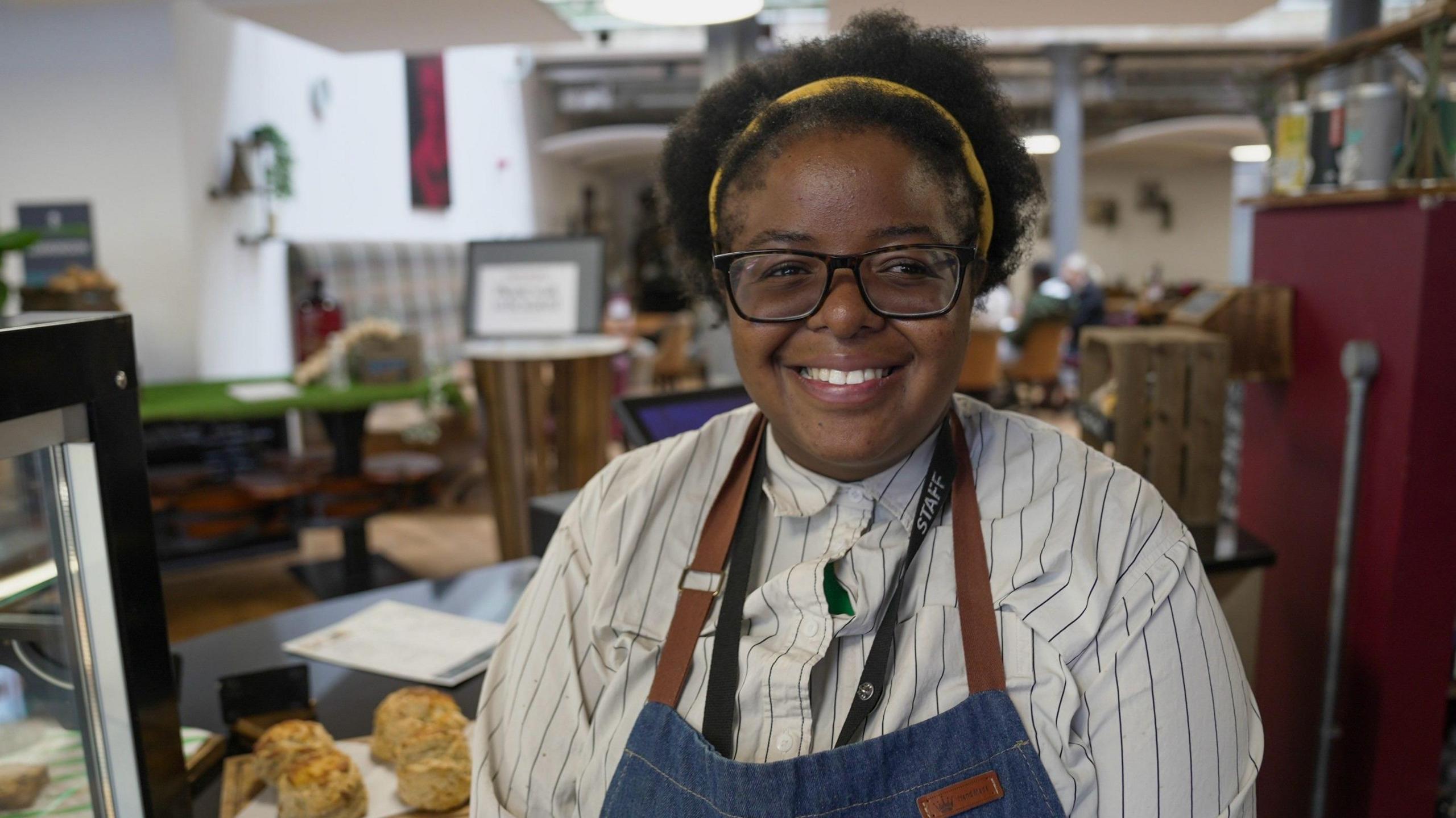 Chachomwe smiles into the camera from behind the counter of the cafe she is working at as part of her apprenticeship. She is wearing a blue denim apron over a white striped shirt and has on glasses and a yellow headband.