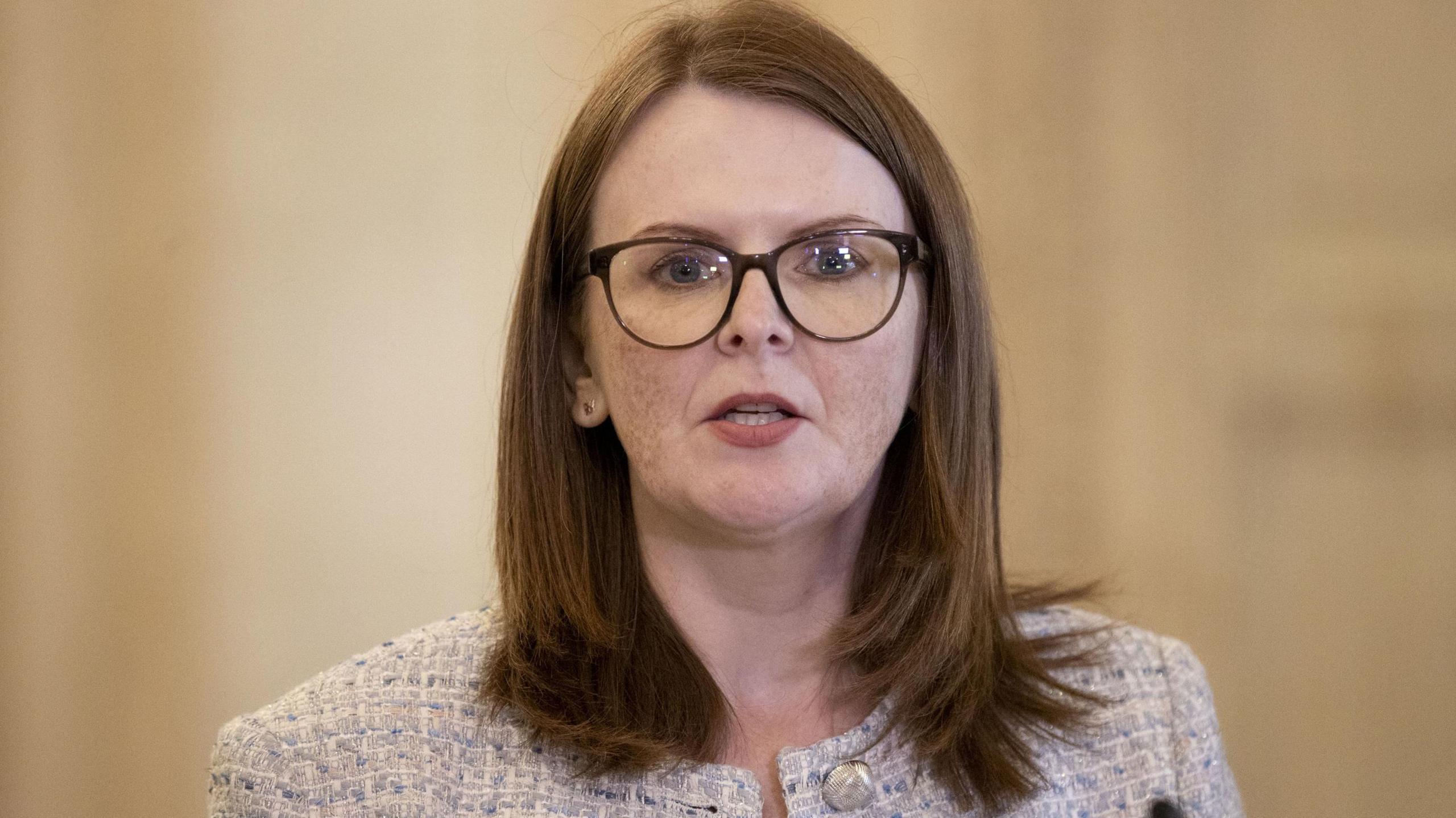 Caoimhe Archibald in white tweed jacket and black top. She has red hair and brown glasses.