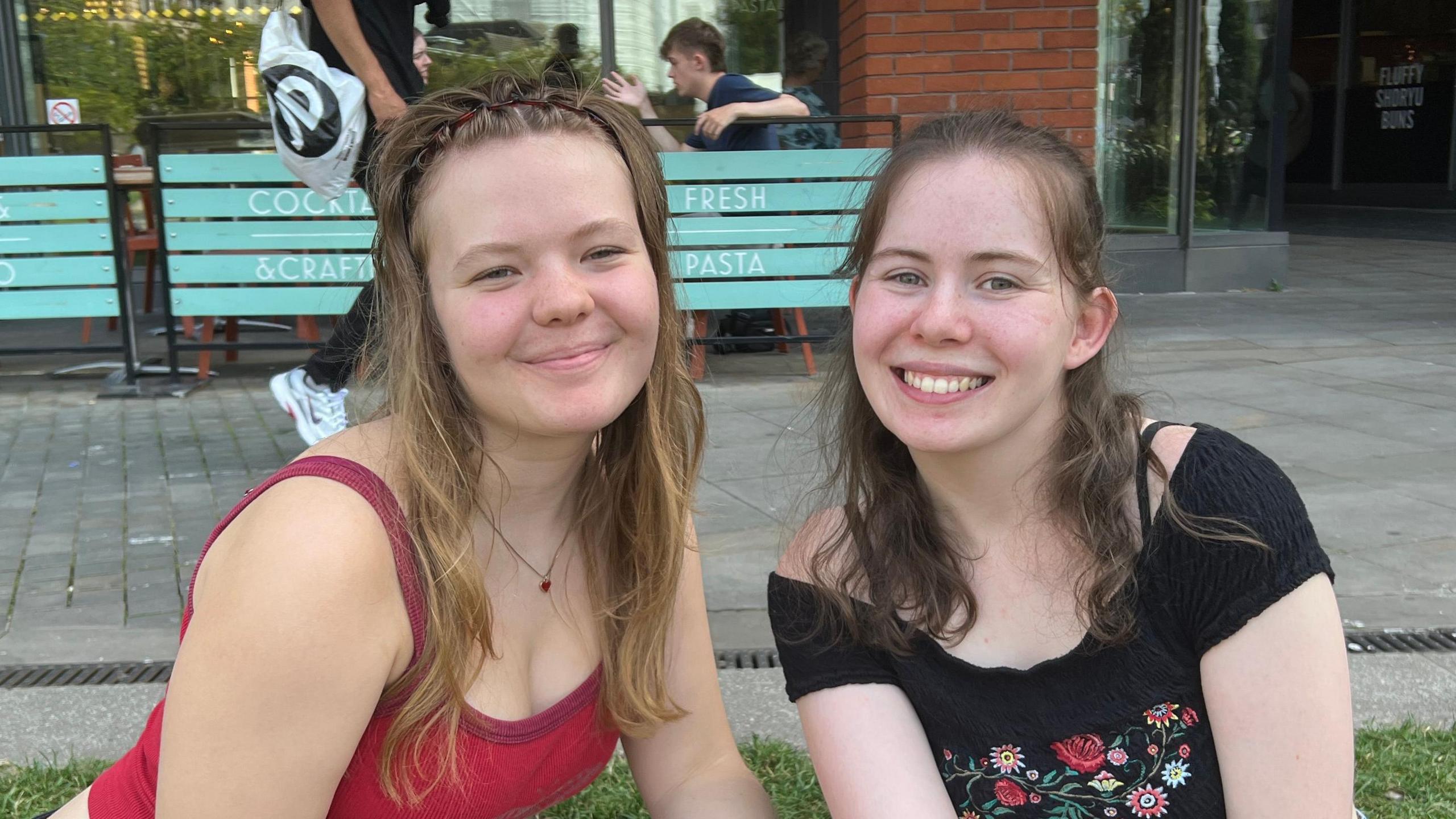 Ms Kerr and Ms Keane are pictured smiling together while sitting on the grass outside. Ms Kerr has long blonde hair and is wearing a headband as well as a red top and a necklace. Ms Keane has long brown hair and is wearing an off-the-shoulder black top with floral detailing.