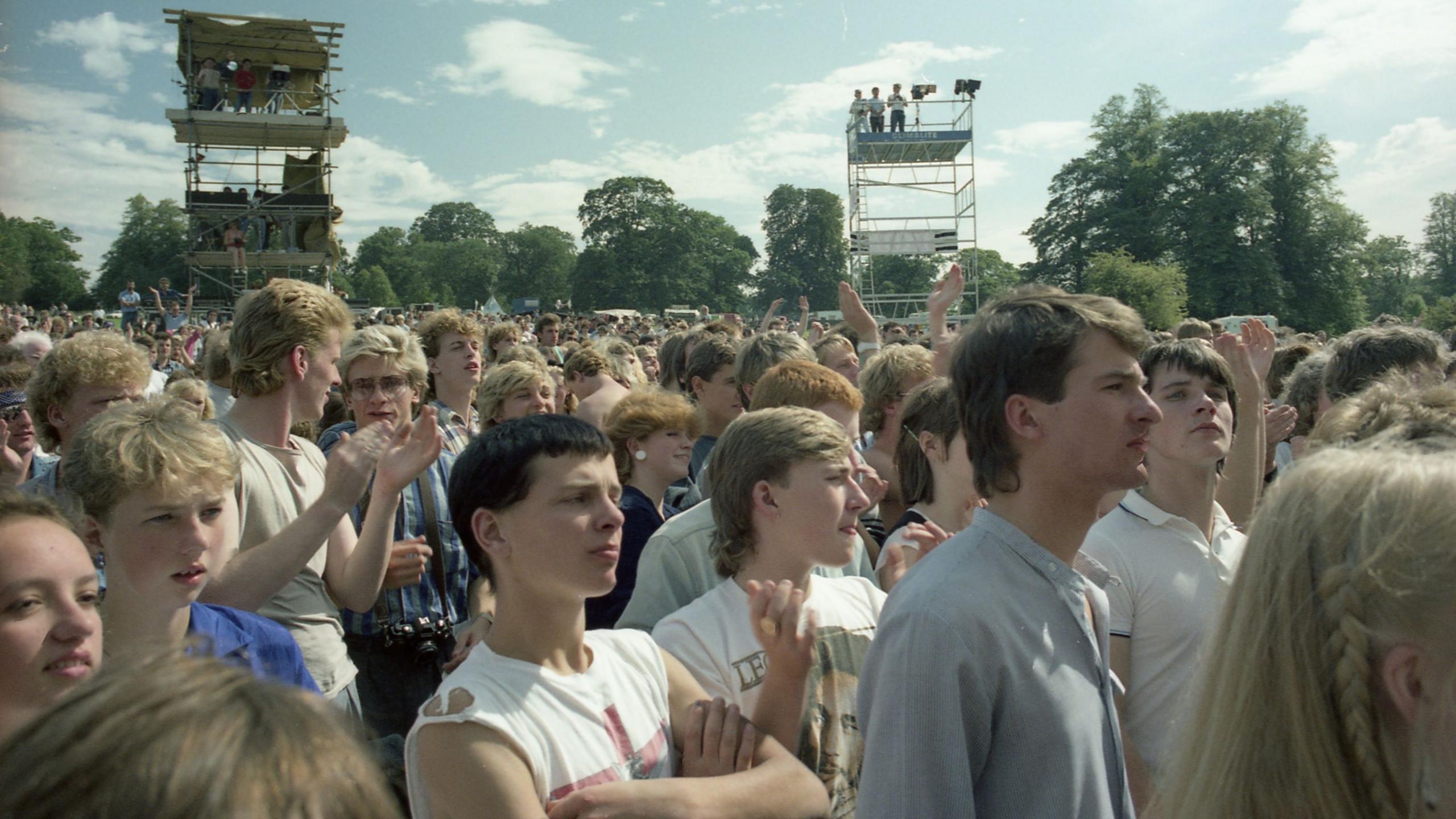 A colour shot of a large crowd of people, mostly young in their teens and 20s, with 1980s clothing and hairstyles. It is an outdoor concert in daylight, and they are all looking towards the stage which is out of picture to the right of frame. Behind the crowd in the background are two scaffolding towers, and a line of trees.