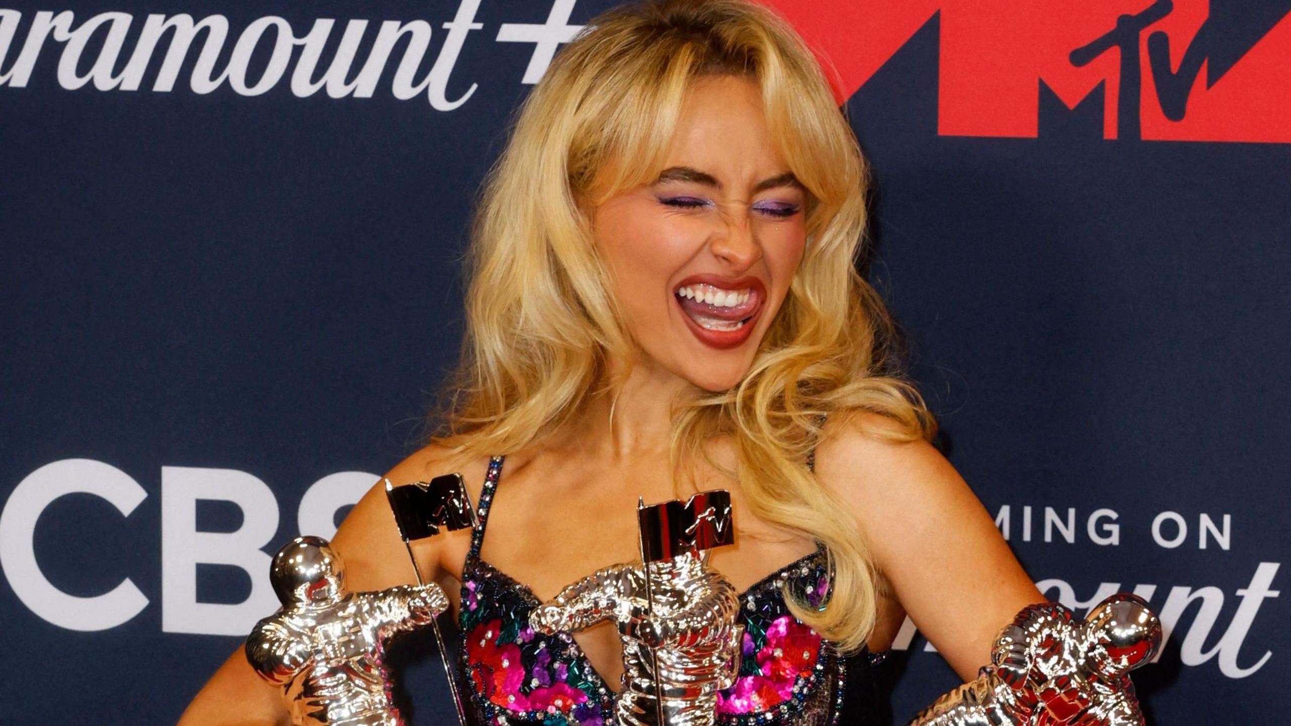 Popstar Sabrina Carpenter pulling a silly face as she holds awards at a red carpet ceremony