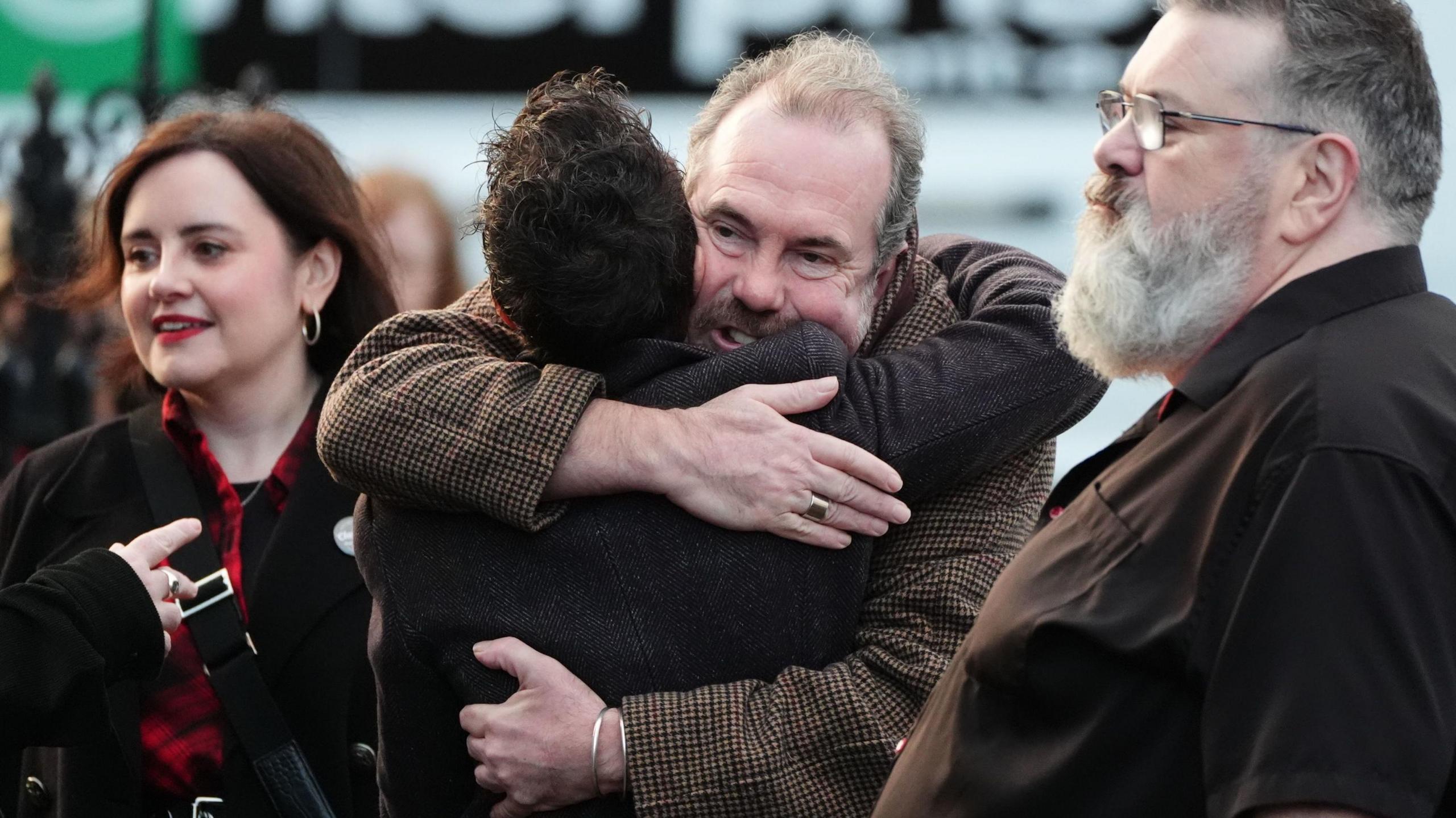 Gavin Mitchell hugs a man in the middle of shot, at the right is a large man with grey heair and glasses and  a younger woman with red lipstick on the left. All are wearing dark colours.