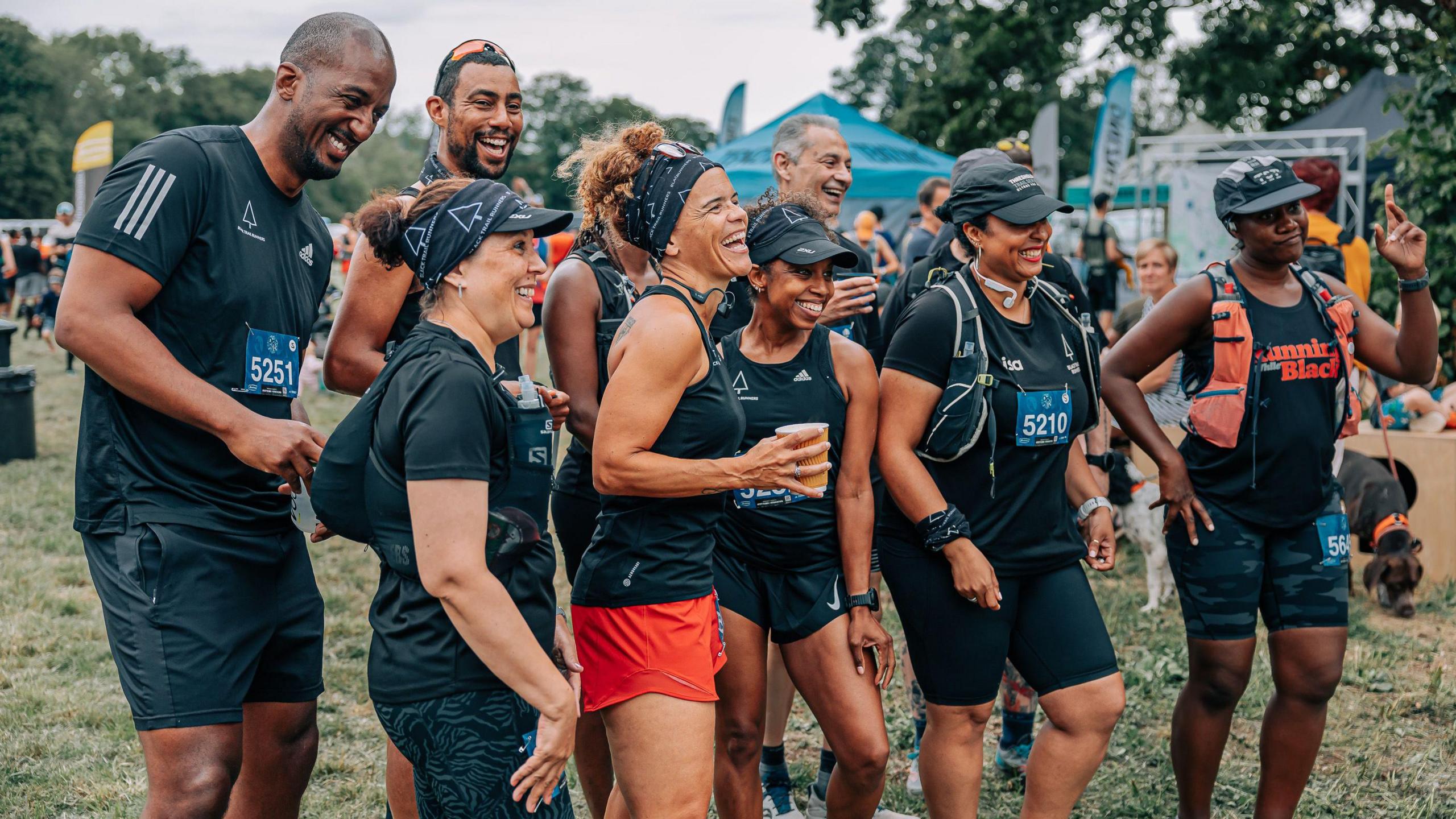 Sabrina Pace-Humphreys with a group of runners all smiling for a camera off to the right. They are all in black running t-shirts and shorts, with Sabrina wearing a black running vest and orange shorts and holding a cup of drink. 