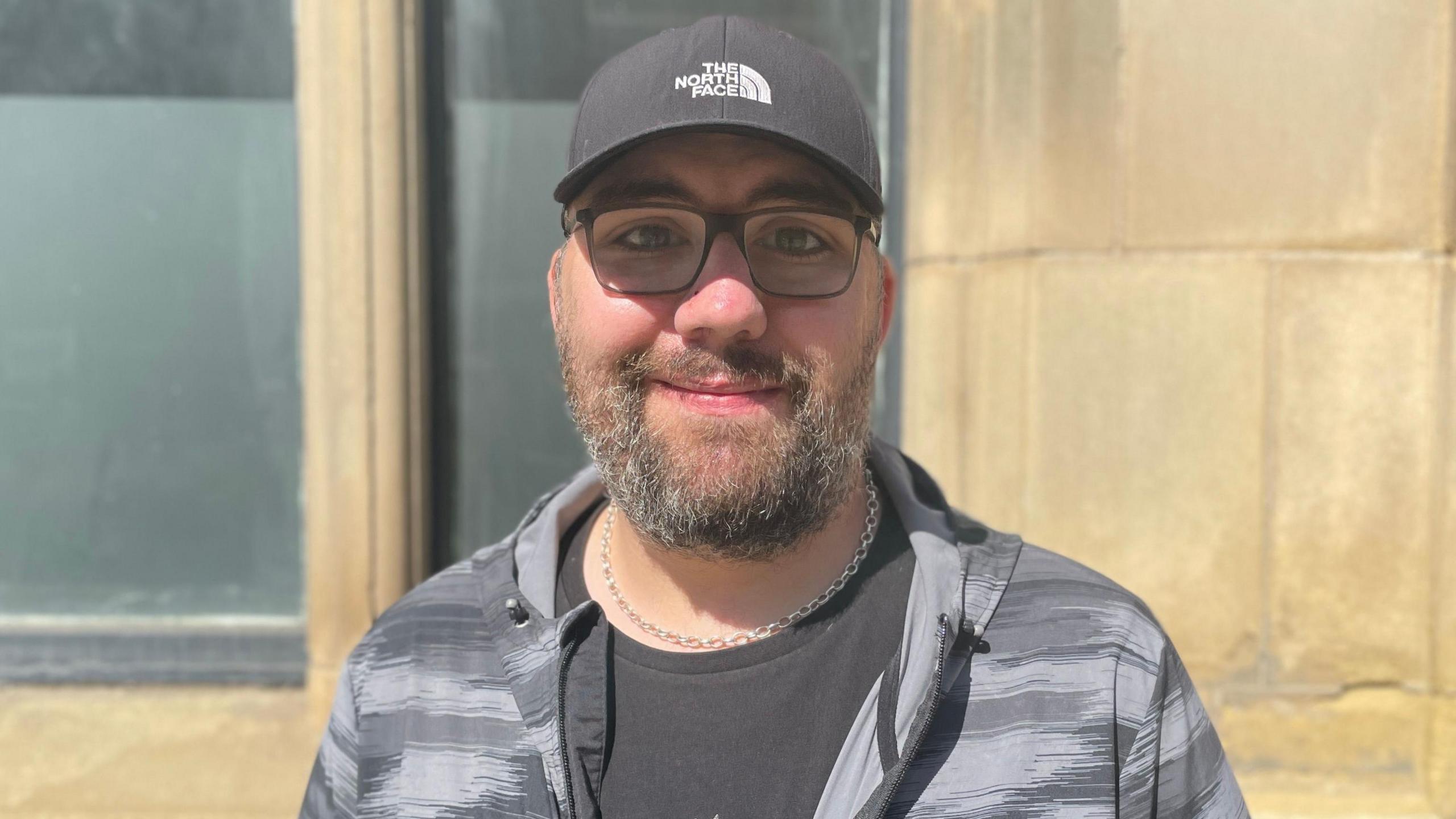 Former homeless man Andy wearing glasses and a black cap smiles at the camera. He has a beard and is wearing a black and grey jacket.