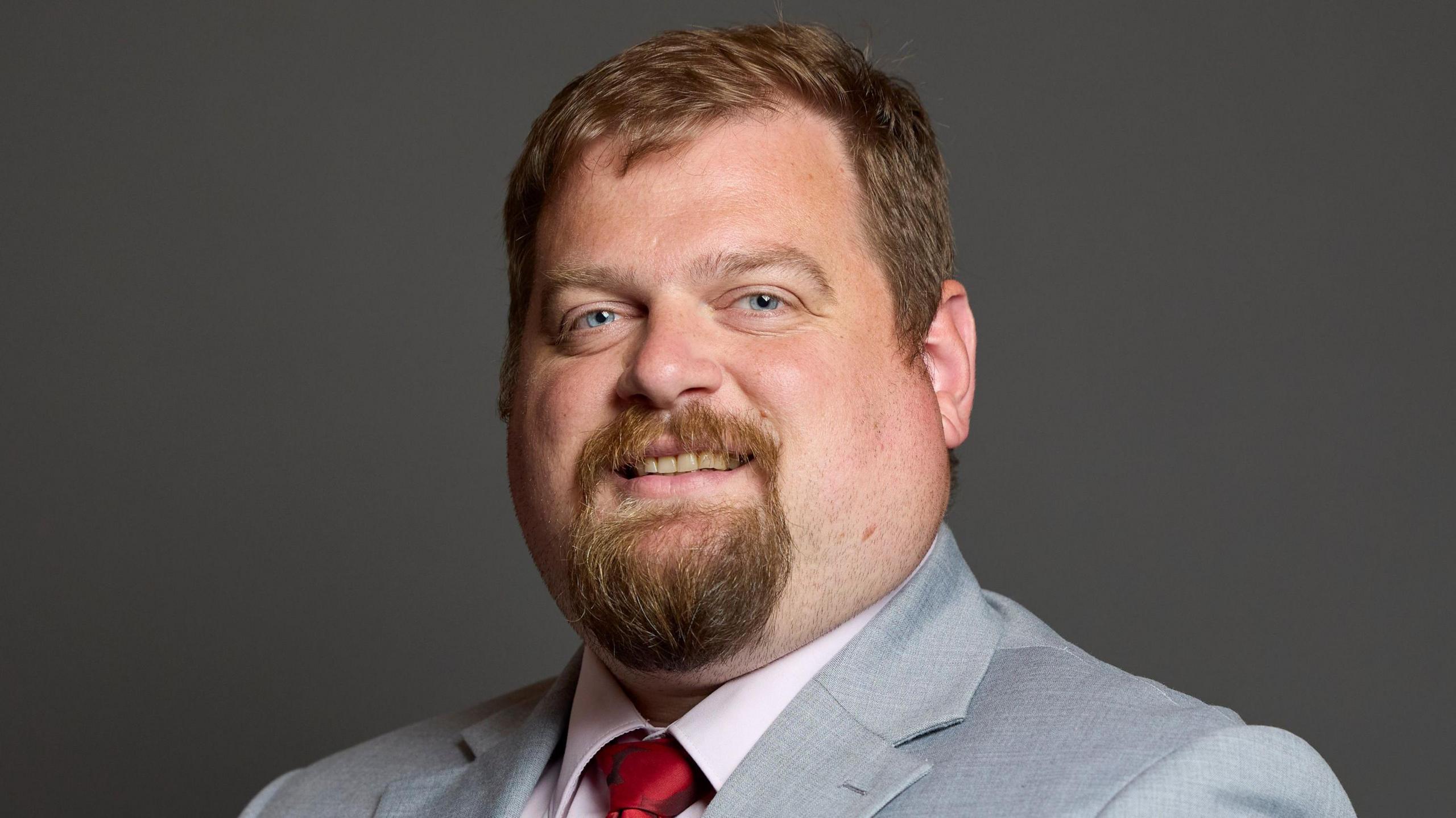 A man with short brown hair and a beard, and wearing a grey suit with a red tie, is smiling at the camera.