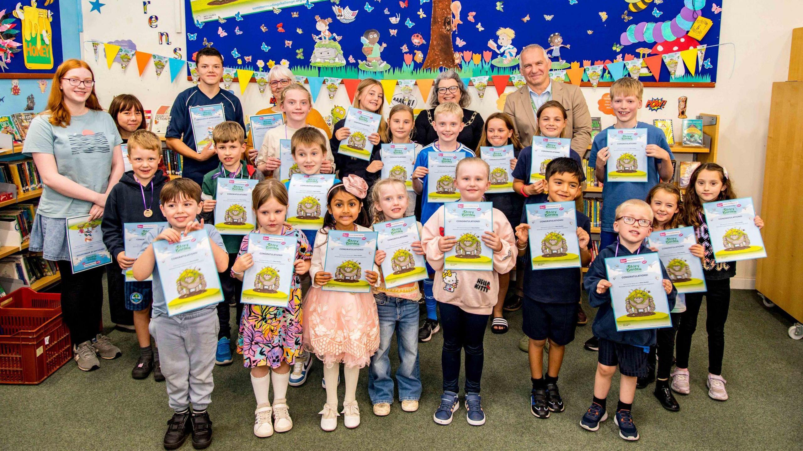 A group of primary school children and a handful of teachers line up in three rows as they face the camera. They are holding up certificates with yellow emblems on them that they were given at the end of a summer holiday reading challenge.