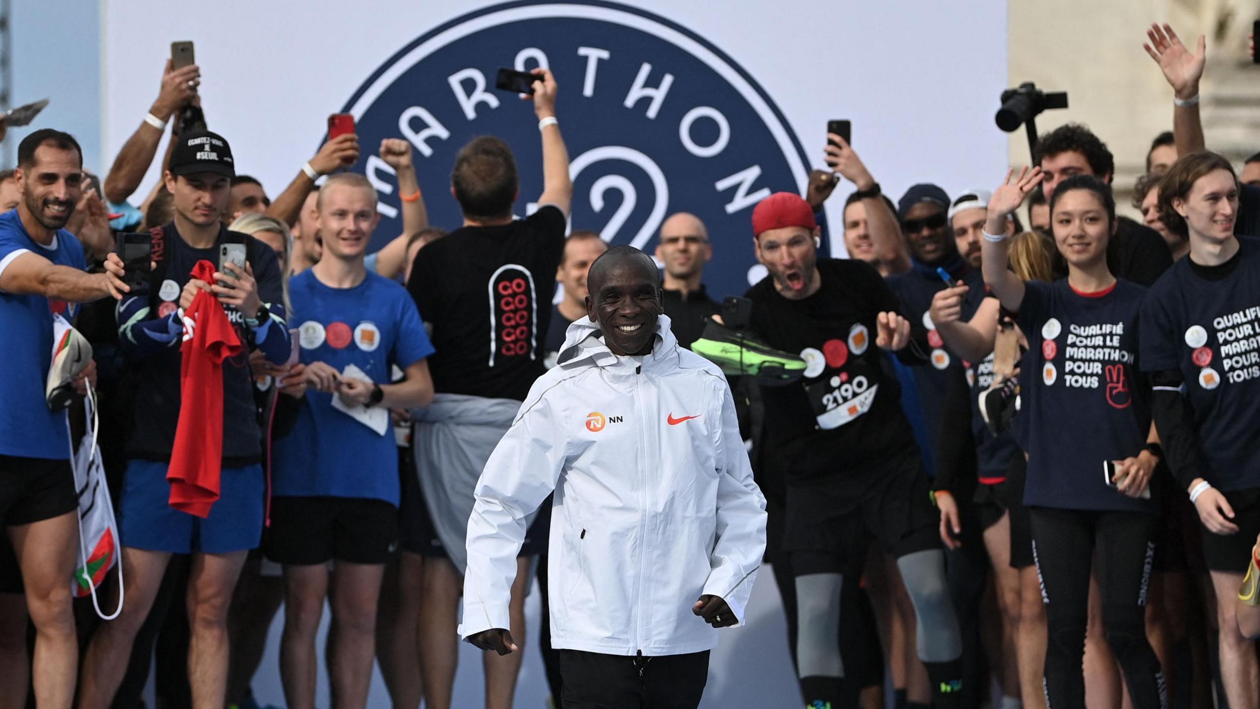 Eliud Kipchoge in white jacket smiles at the camera as members of the public due to run a race with him smile and point in the background