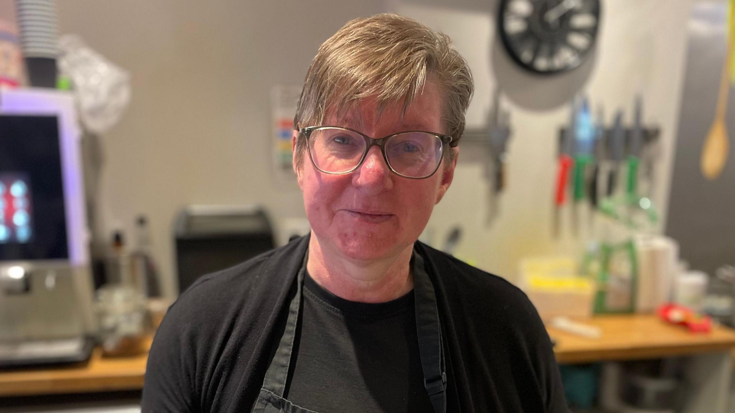 Sandra has short brown hair and is wearing glasses. She is wearing a black top.