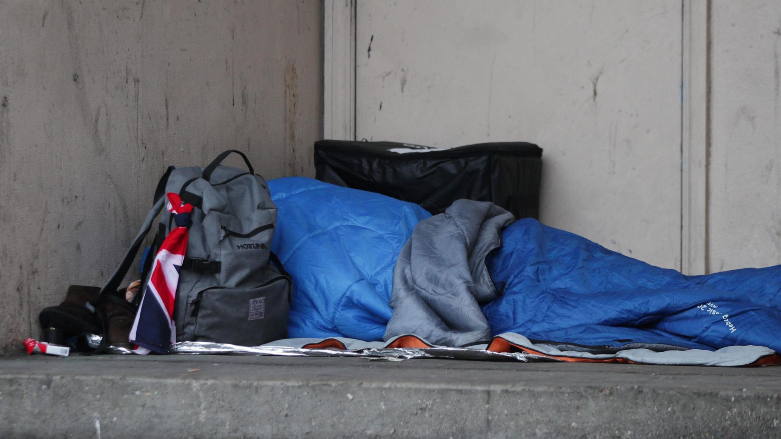 A blue sleeping bag is seen in an alcove, alongside is a grey rucksack which appears to have a Union Jack flag tied to it. There are various other items nearby, including a packet of cigarettes.