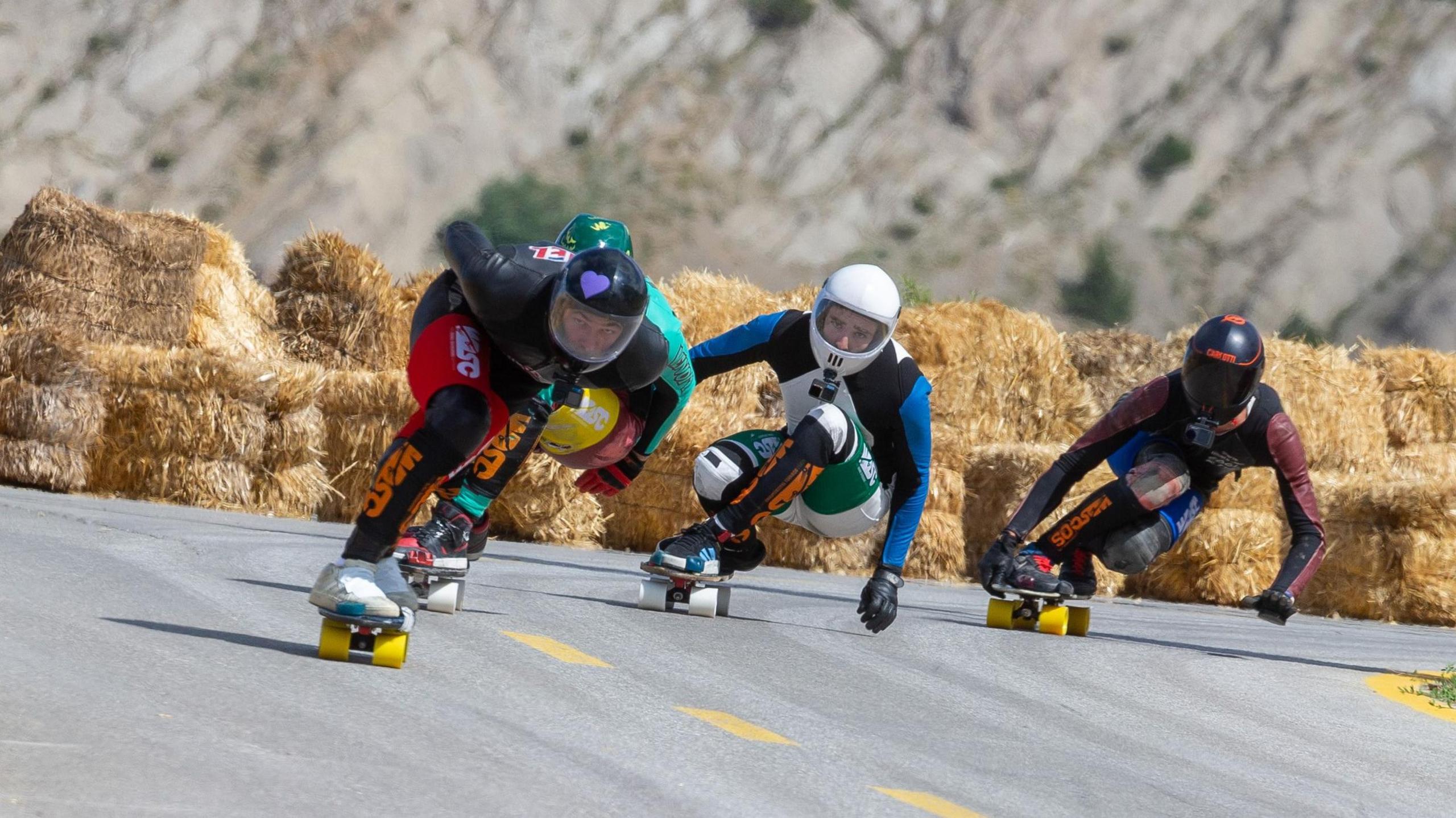Four people in brightly coloured leathers and helmets on skateboards bend down on a grey road. Yellow hay bales lining the course can be seen in the background, with a rock cliff face behind them.
