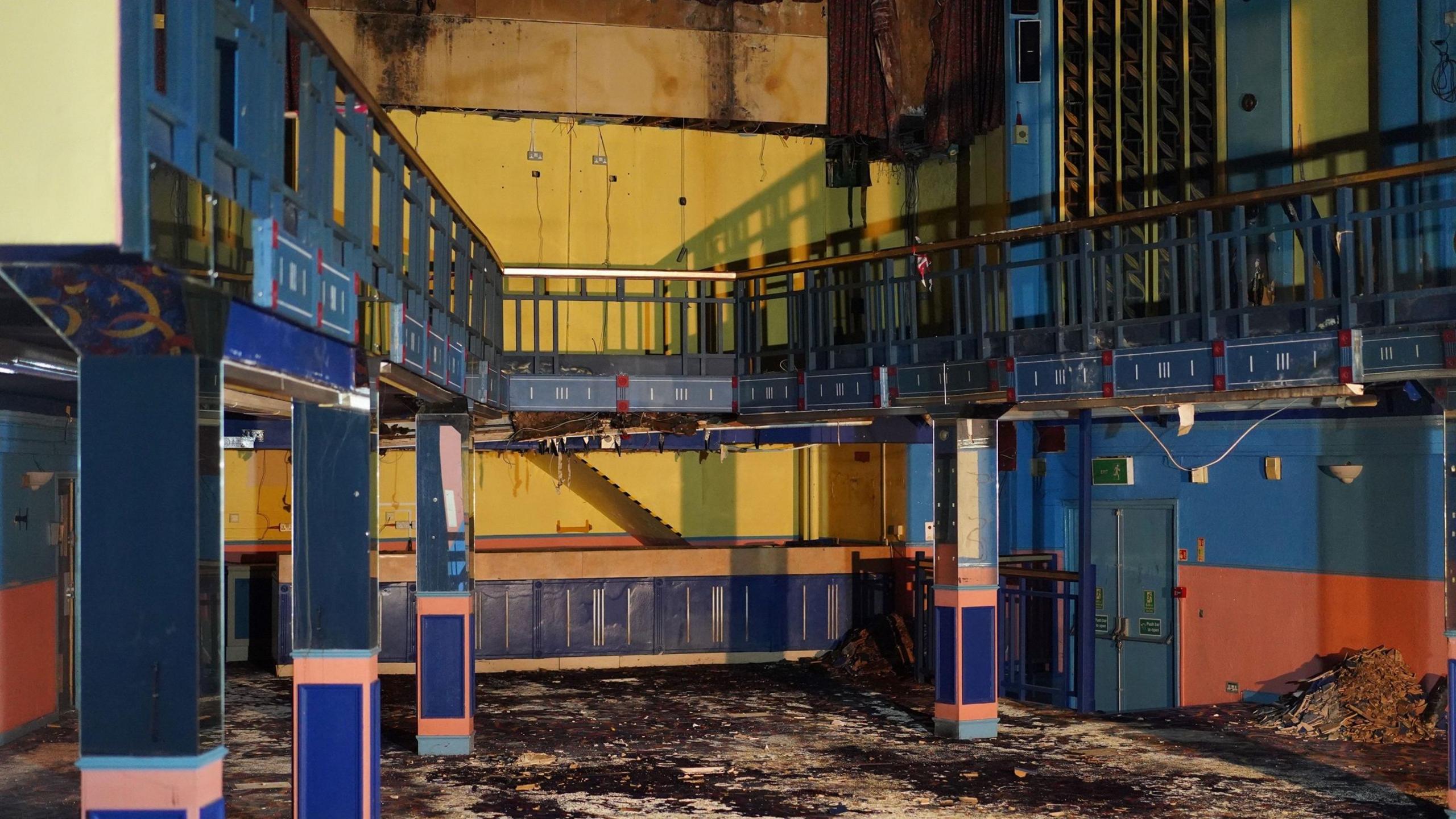 A derelict open space inside the building. There is a mezzanine painted in blue and in front is a yellow wall that once had the cinema screen. The carpet is discoloured and dirty, with a pile of rubble in the corner.
