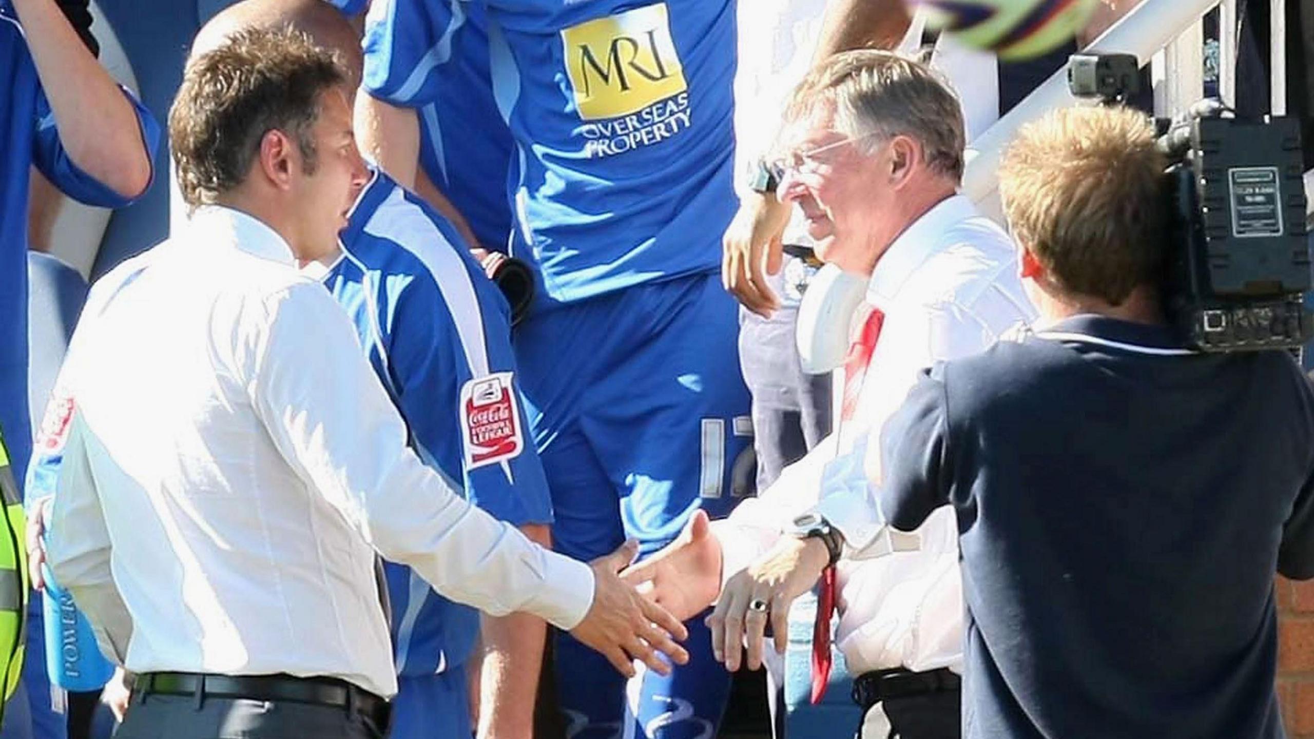 Darren Ferguson, dressed in a white shirt, shakes hands with his father, Sir Alex Ferguson, following a game between Peterborough United and Manchester United in August 2007, with a cameraman filming the moment stood nearby