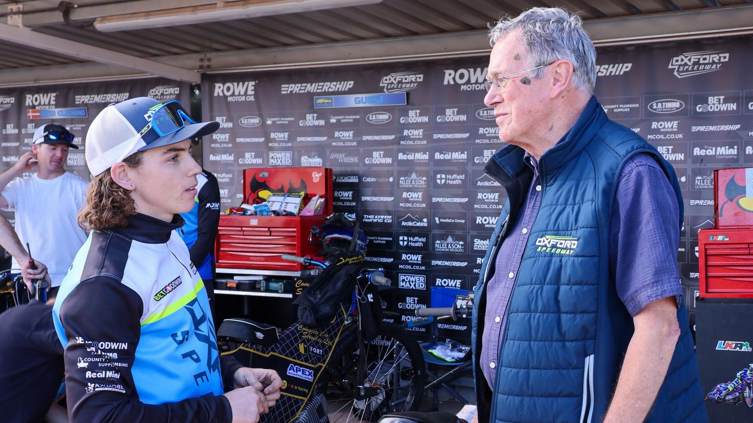 Ian Wagstaff has grey hair and glasses, and is talking to a speedway rider - who is wearing a cap and has medium length brown hair.