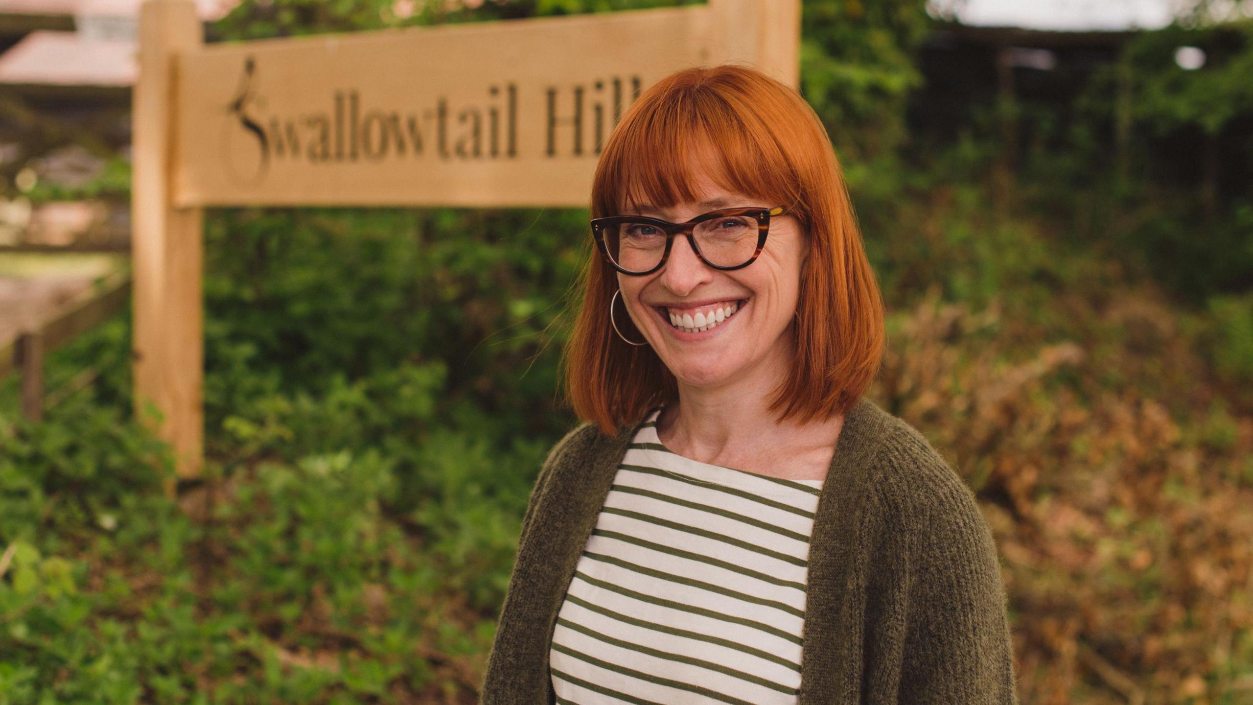 Sarah Broadbent is wearing a white and brown striped top, a green cardigan and dark framed glasses. She is standing in front of a sign which says Swallowtail Hill